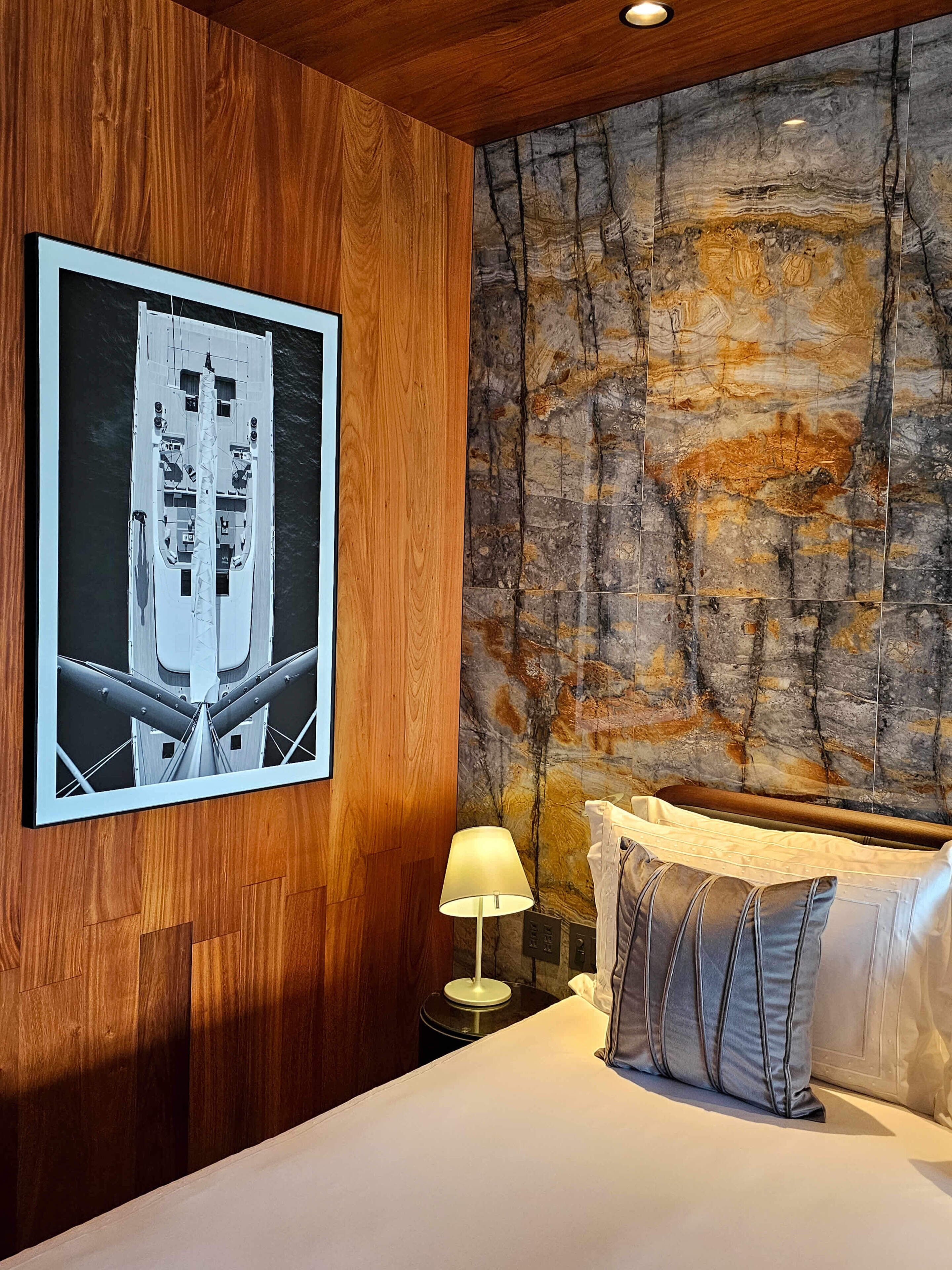 A hotel room with a wooden panel wall, a large abstract artwork, a bed with pillows, and a nightstand with a lamp.