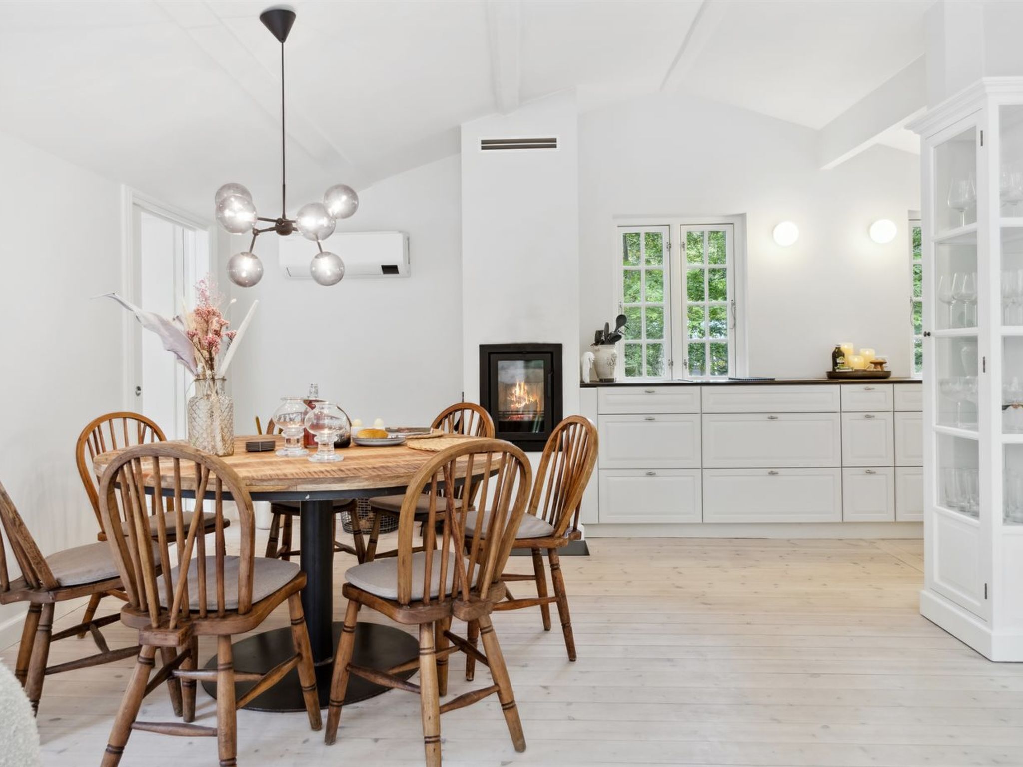 A modern kitchen with white cabinetry, a dining table with wooden chairs, a built-in stove, and a view of greenery through the windows.