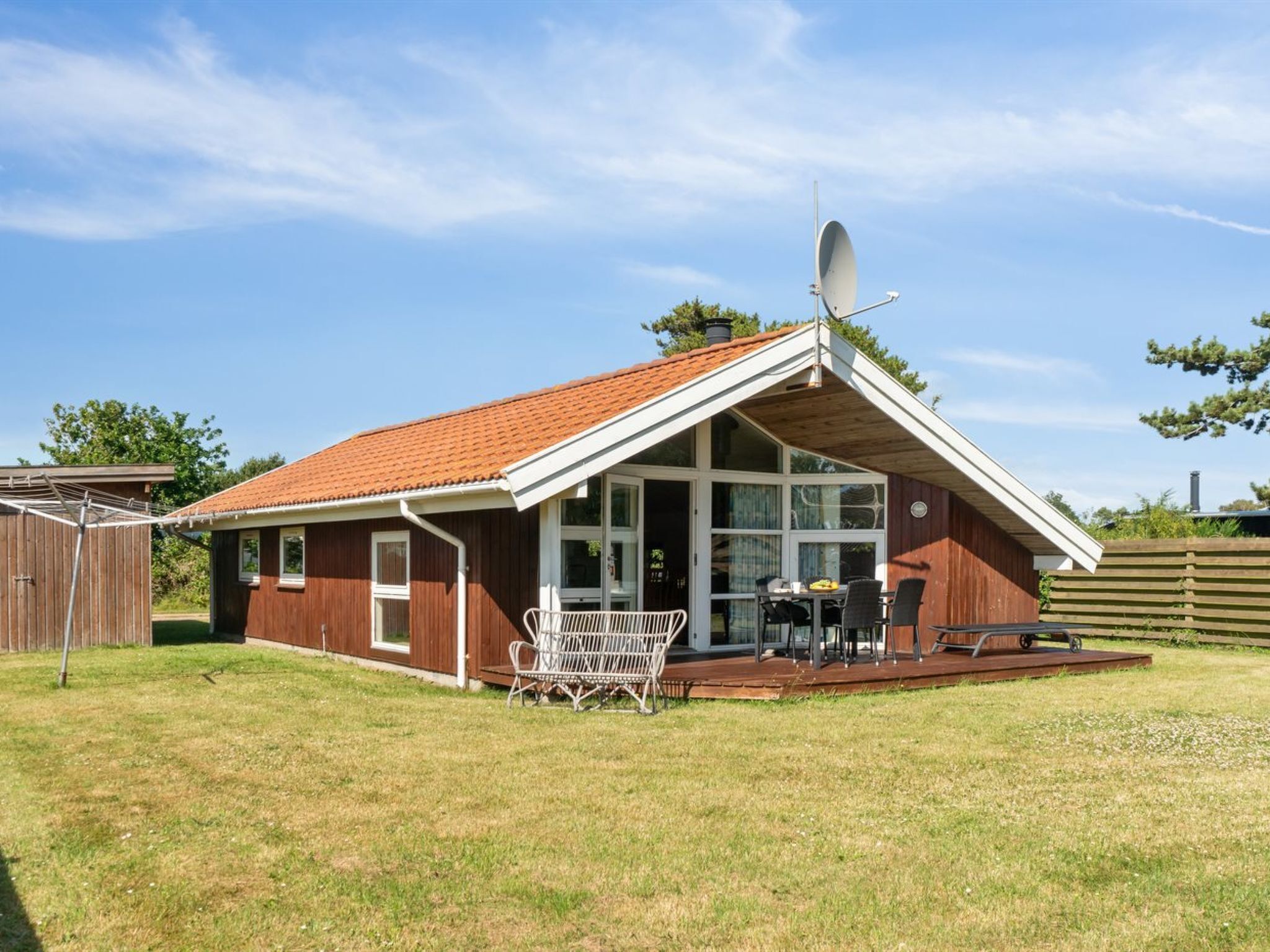 Une maison de plain-pied avec un toit recouvert de tuiles rouges, une antenne parabolique et un patio couvert comprenant une table et des chaises.