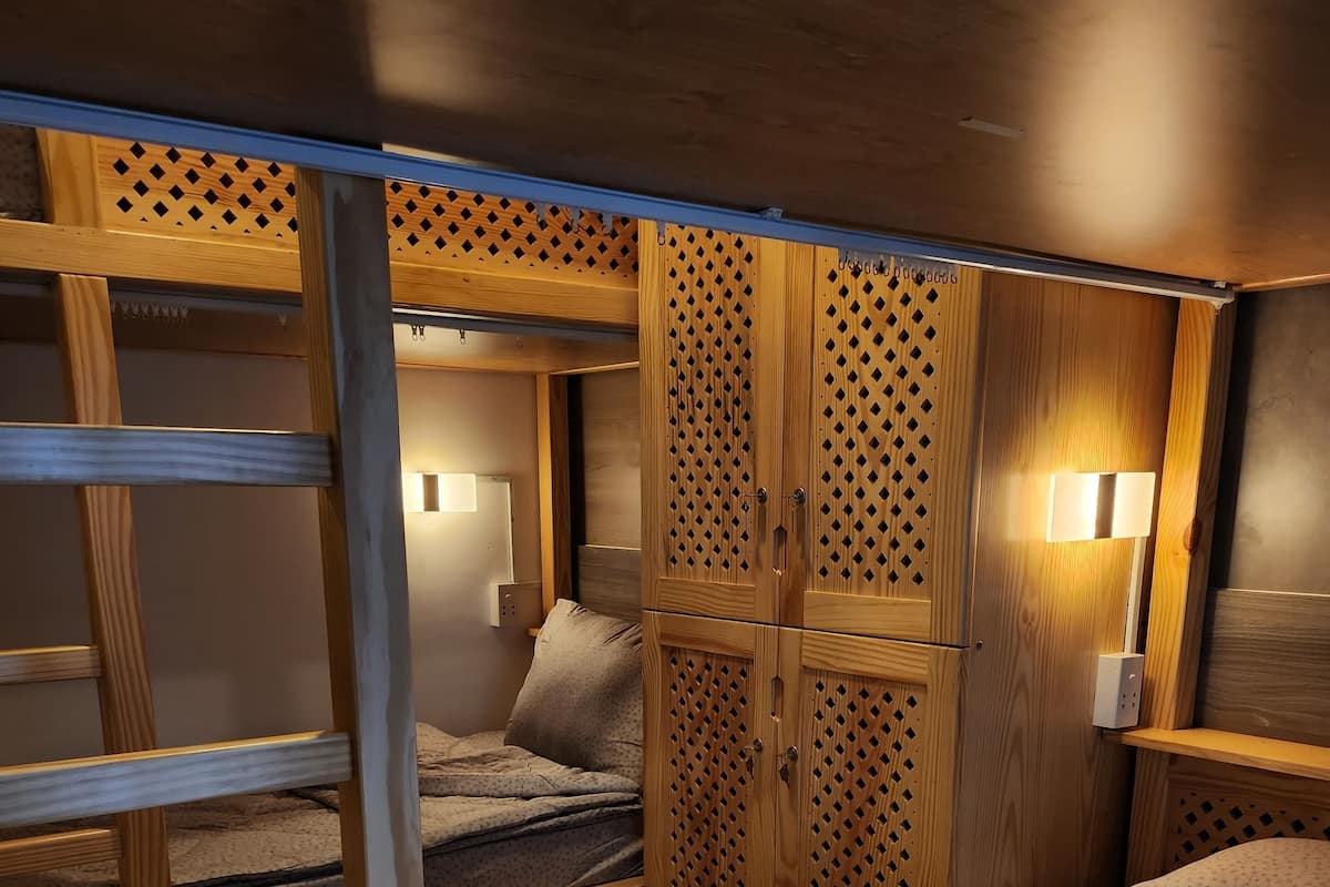 A compact, well-lit room with a bunk bed, wooden wardrobe with a lattice design, and a bedside table with a lamp.
