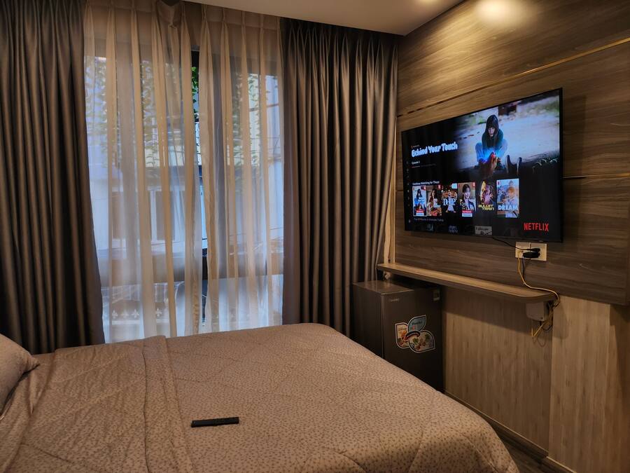 Una habitación de hotel con cama, un televisor montado en la pared mostrando contenido de Netflix y una ventana con cortinas traslúcidas.