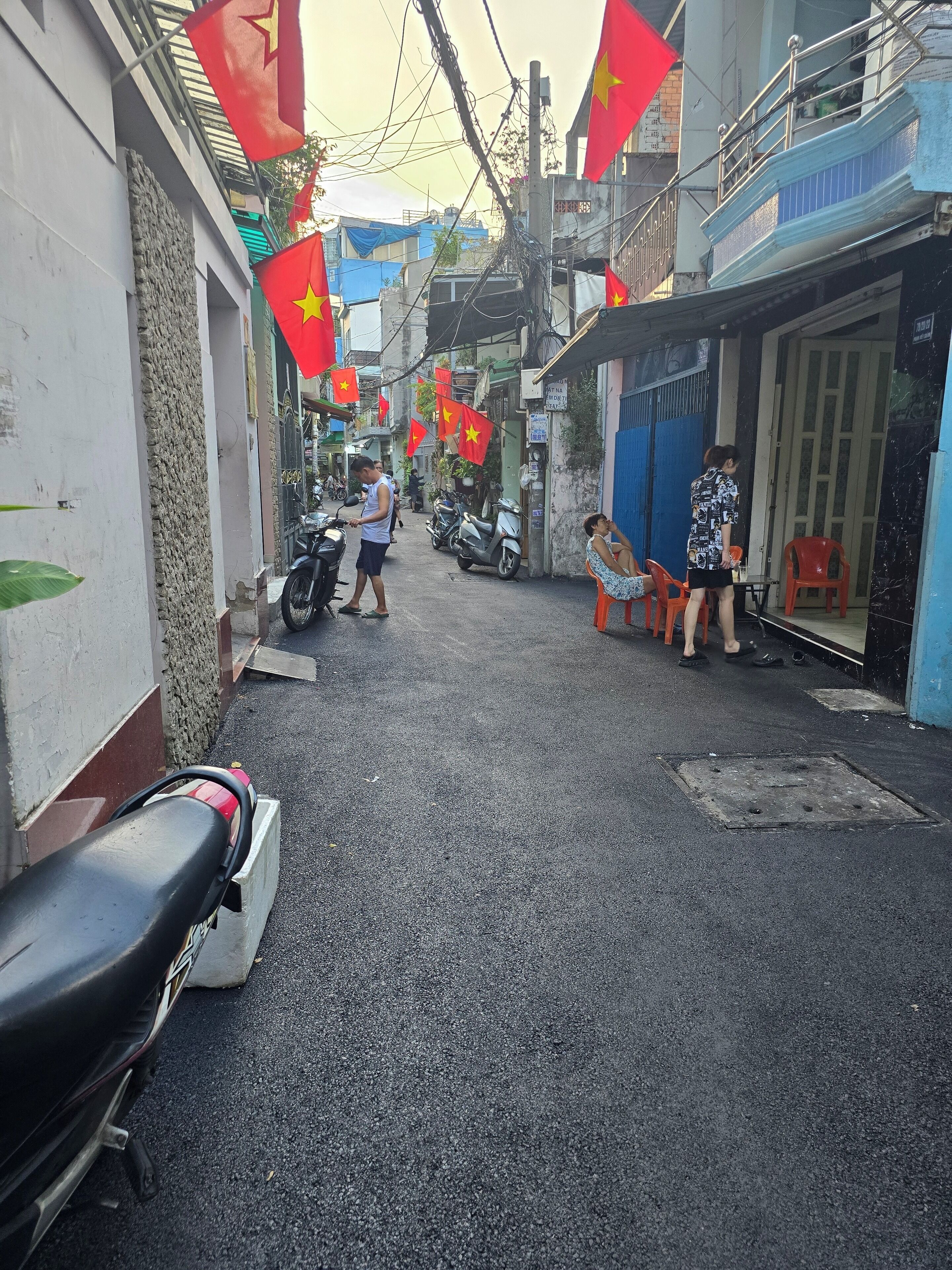 Une rue étroite, bordée de scooters et de motos garés, où quelques personnes se promènent, et où flottent des drapeaux rouges.
