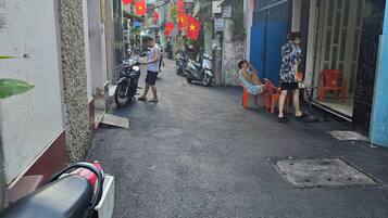 A narrow street with parked scooters and motorcycles, a few people walking, and red flags hanging.