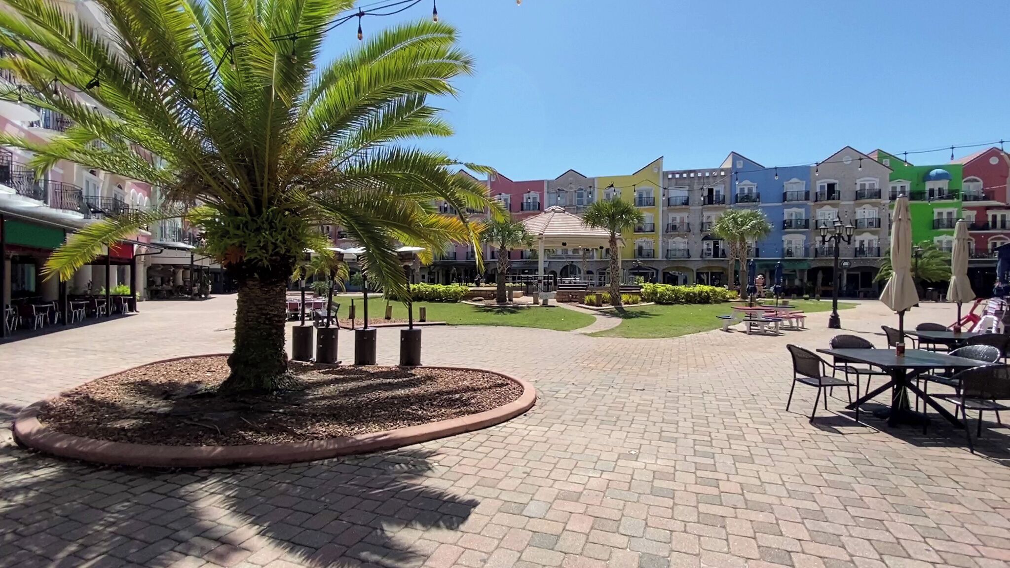 A courtyard with palm trees, outdoor seating, and colorful buildings in the background.