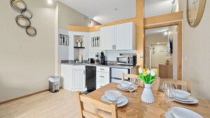 Una cucina moderna con mobili bianchi, un tavolo da pranzo in legno apparecchiato con piatti e bicchieri, e un piccolo cestino.
