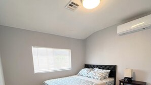 Une chambre à coucher comprenant un lit, une table de chevet, une fenêtre avec des stores, un luminaire au plafond et un appareil de climatisation.
