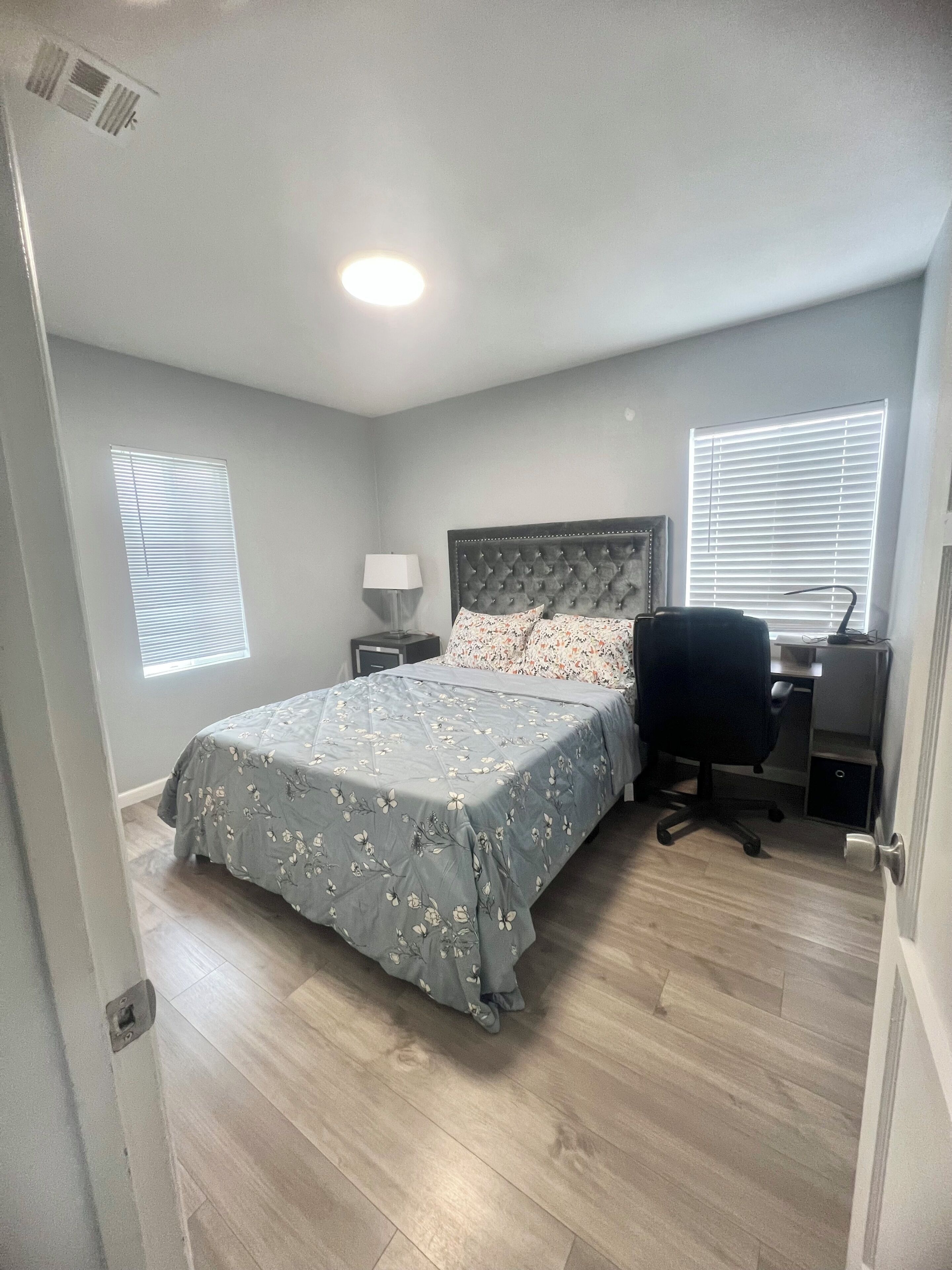 Une chambre à coucher comprenant un lit, un bureau, une chaise, une lampe et deux fenêtres équipées de stores.