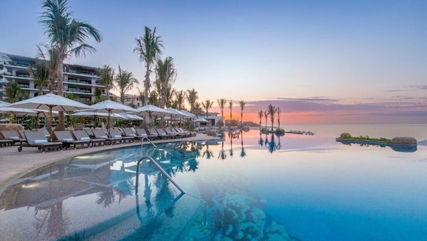 An infinity pool with lounge chairs and umbrellas, surrounded by palm trees and a hotel building. -  VILLA LA VALENCIA, LOS CABOS, TWO BEDROOM OCT28-NOV5TH (San José del Cabo)