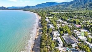 An aerial view of a coastal town with a beach, residential buildings, and a mountainous backdrop. - The Santana Collection TWO - Luxury Villa Retreat in Palm Cove with Private Pool (Palm Cove)