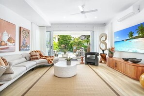 Ein geräumiges Wohnzimmer mit großem Fernseher, einer Couch, einem Couchtisch und Blick auf einen tropischen Strand durch die Glastür.