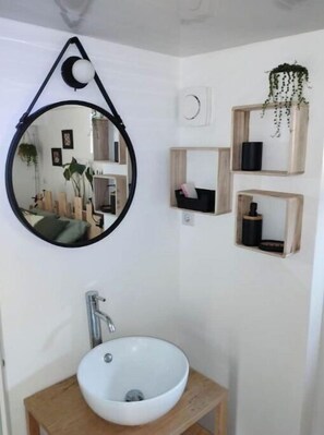 Une salle de bain avec un miroir rond, un lavabo blanc et des étagères en bois ornées de plantes et d’articles de toilette.