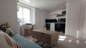 A compact living space with a sofa, a small table, a kitchenette, and a trash bin. - Appartement Proche Gare et Centre-ville D'amiens (Rivery)