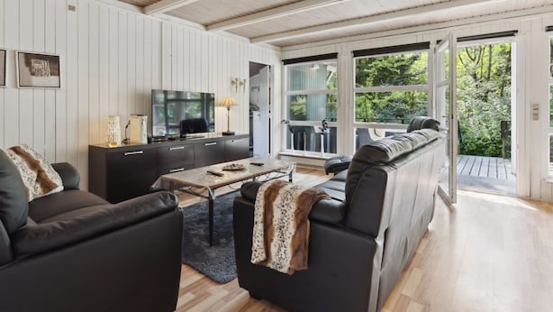 A modern living room with a sofa, a coffee table, and a TV. There is a large window with a view of greenery outside.