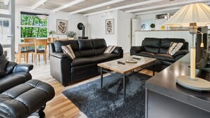 A modern living room with black leather sofas, a coffee table, and a lamp. There is a dining area with wooden furniture and a view of greenery outside.