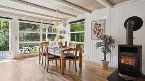 A dining area with a wooden table and chairs, a large window with a view of greenery, and a wood-burning stove.