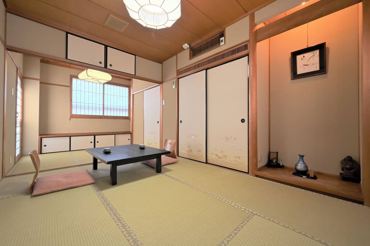Une pièce de style japonais traditionnel, avec un sol recouvert de tatamis, une table basse, un fauteuil rembourré, des portes coulissantes et une étagère ornée d’objets décoratifs.