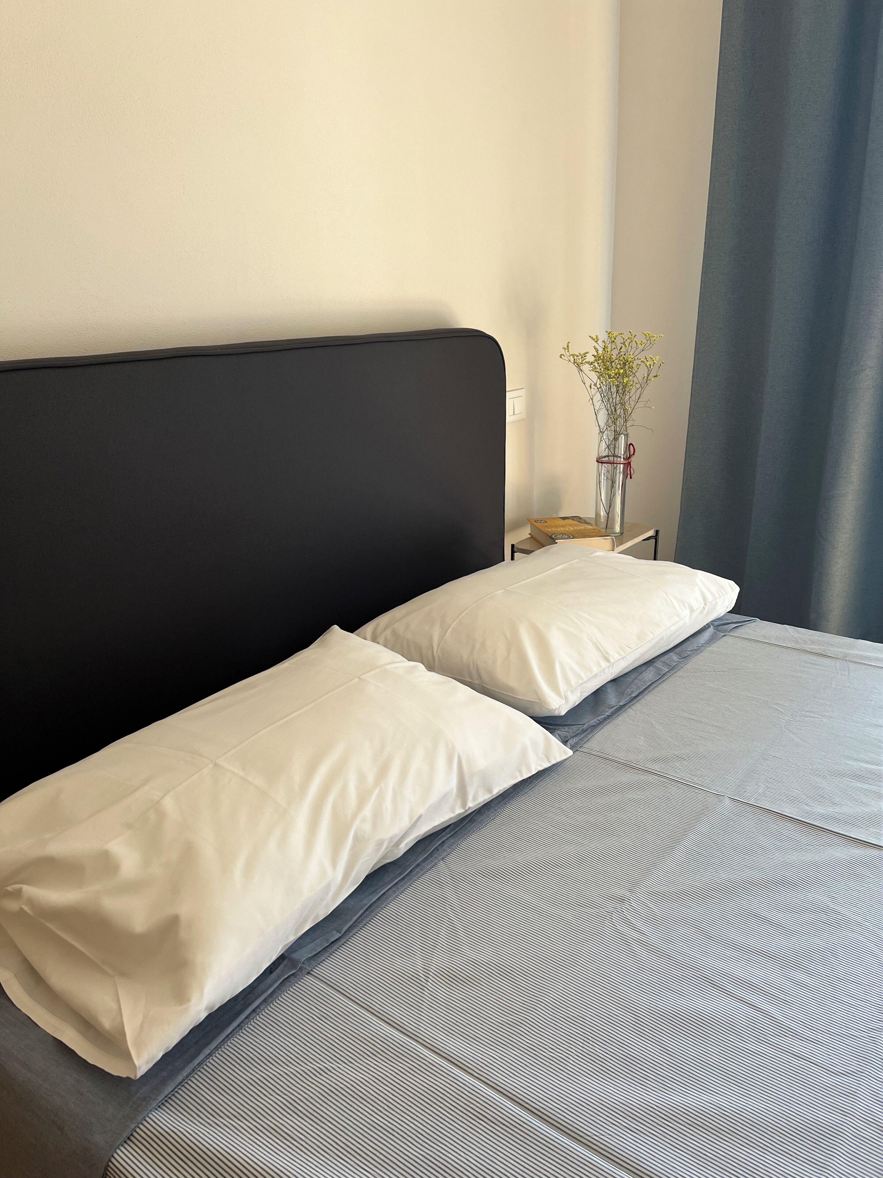 Ein ordentlich bezogenes Bett mit weißen Kissen und einer grauen Tagesdecke, ein kleiner Nachttisch mit Blumen in einer Vase und ein dunkler Vorhang am Fenster.