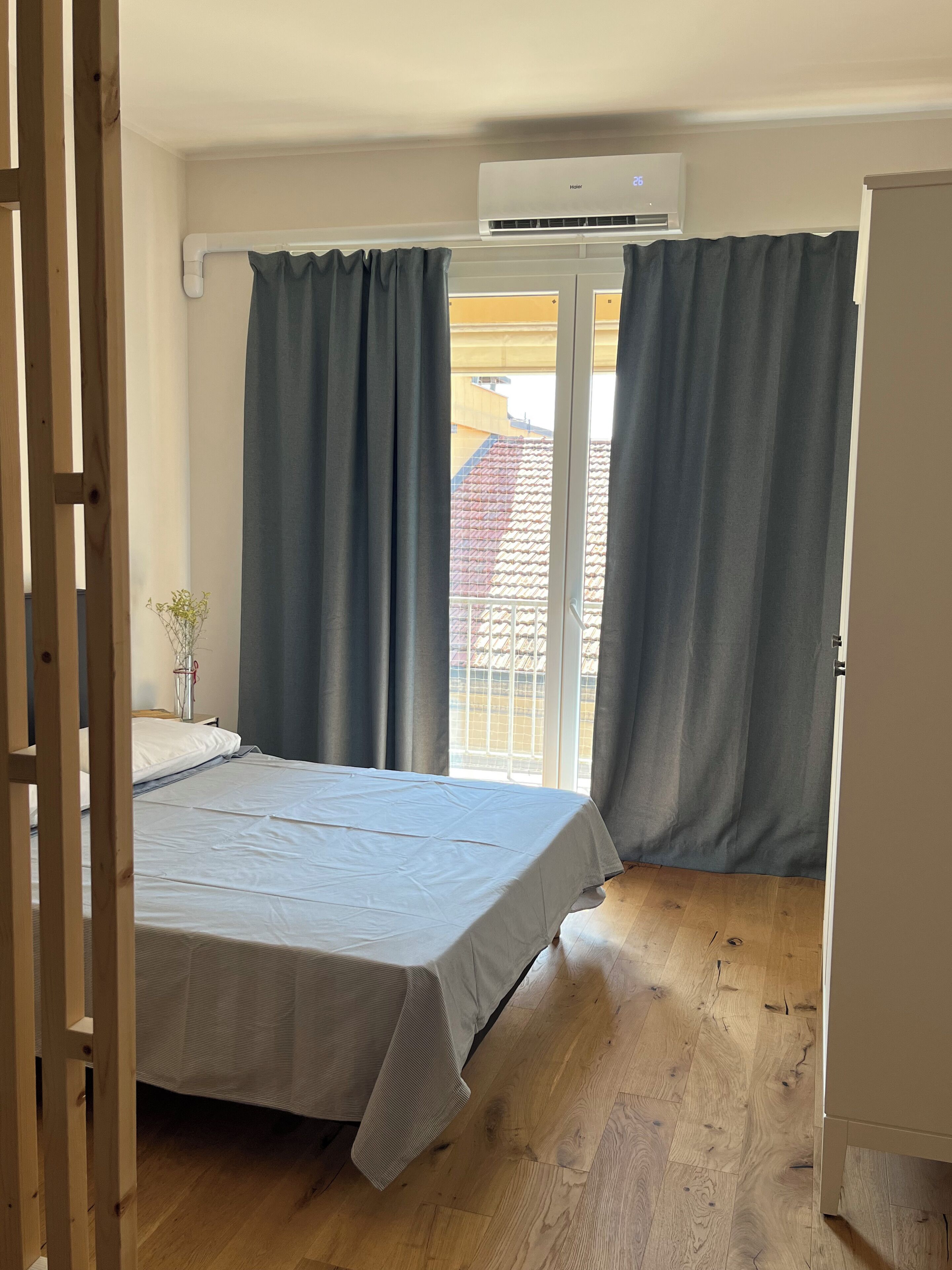 Une chambre avec un lit, une fenêtre avec des rideaux, un appareil de climatisation et une vue sur un bâtiment à l’extérieur.