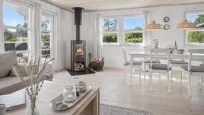 A modern living room with a wood-burning stove, white furniture, and large windows offering a view of the outdoors.