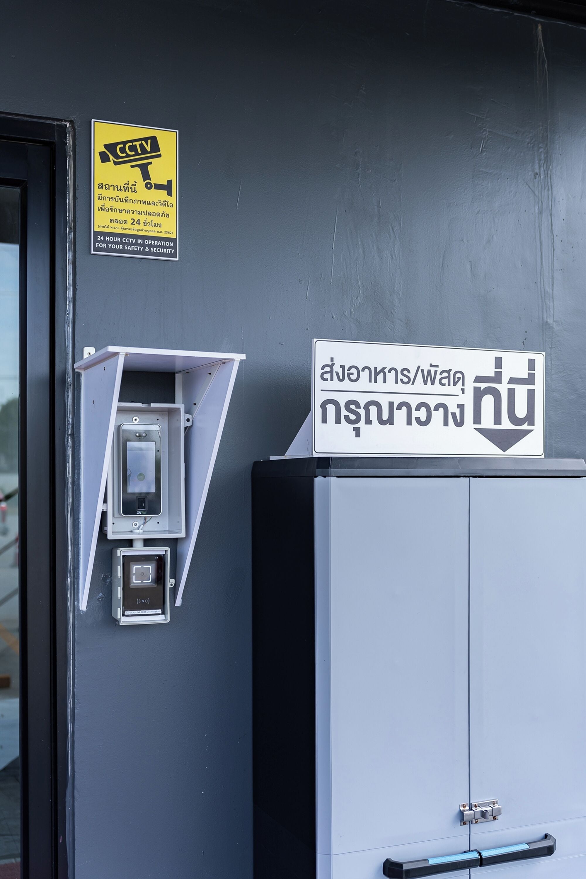Une caméra de surveillance fixée à un mur, un panneau avec du texte en thaï, et une armoire technique dotée d’une porte.