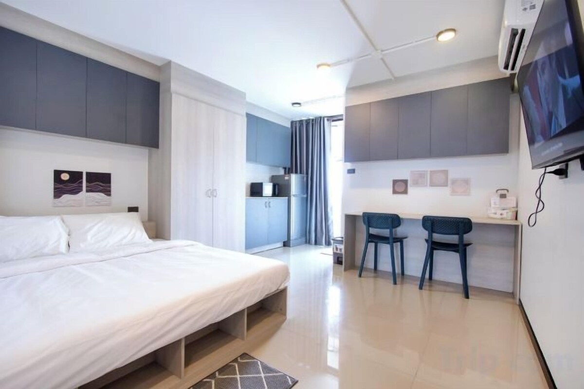 A modern hotel room with a bed, a desk, two chairs, a TV, and a kitchenette.
