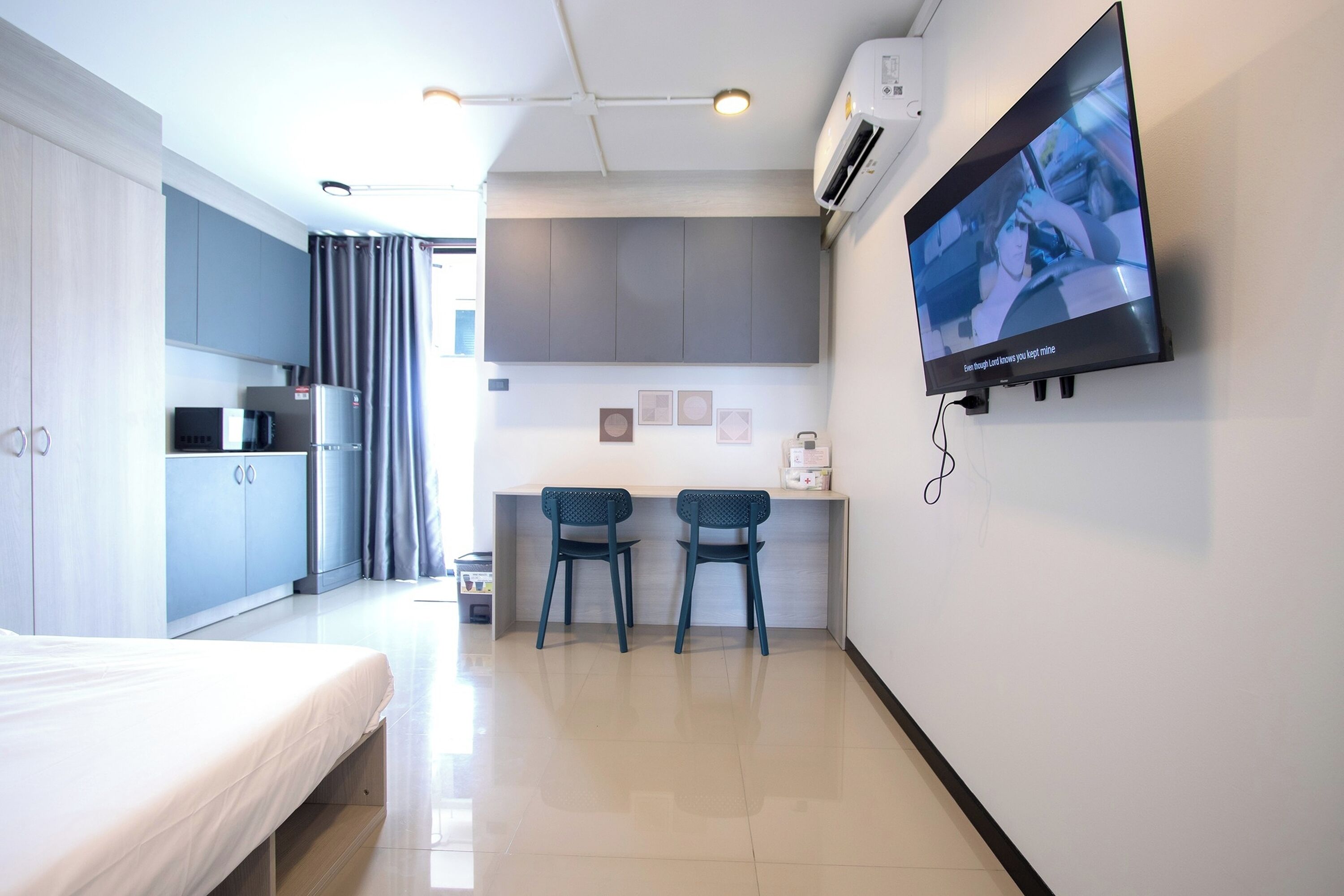 Una habitación de hotel moderna con cama, kitchenette, dos sillas, televisor y aire acondicionado instalado en la pared.