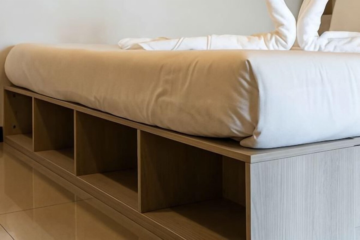 Una cama con colchón y almohadas blancas, una base de madera debajo y una pared de color claro.