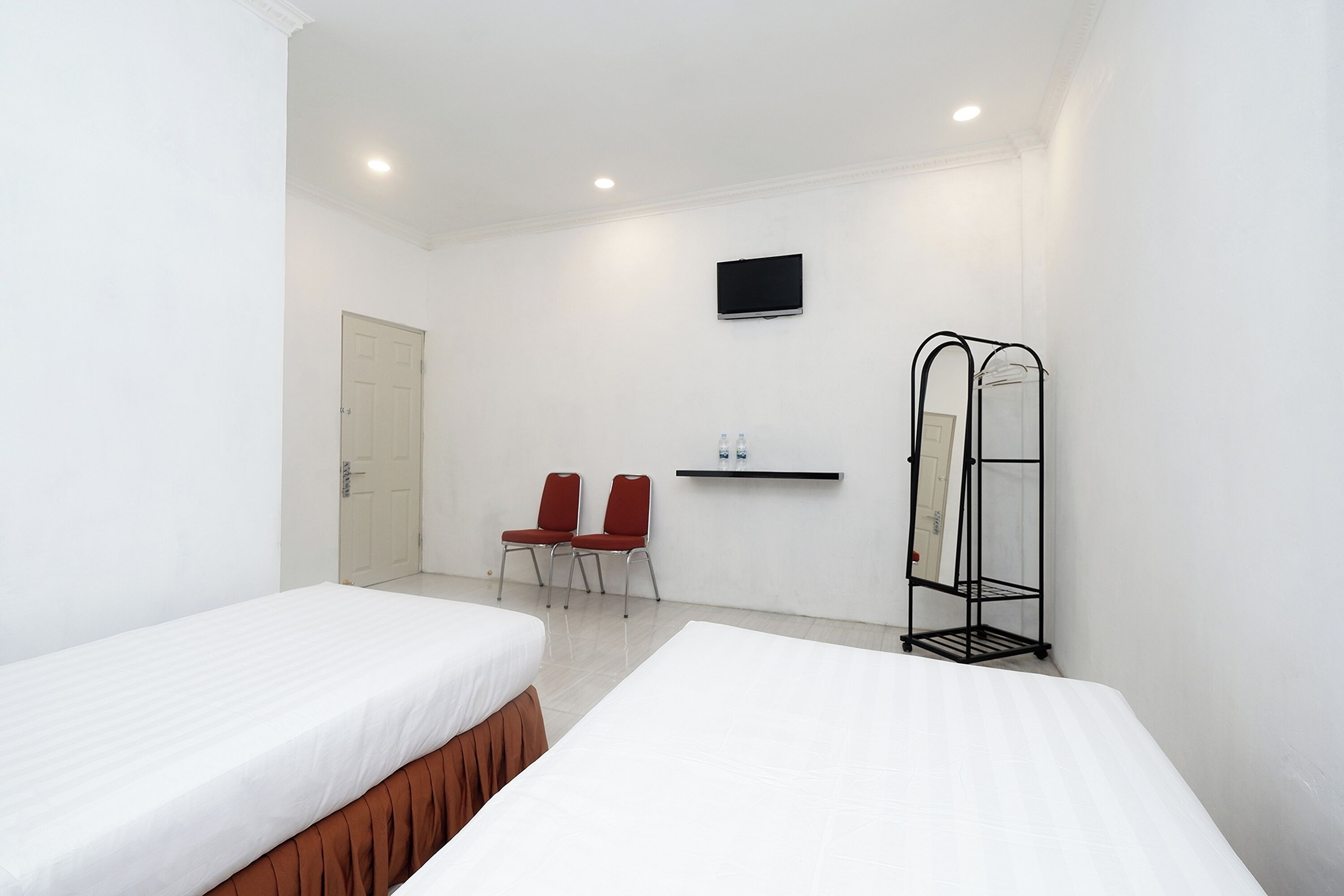 A hotel room with two beds, a TV, a shelf with bottles, and a coat rack.
