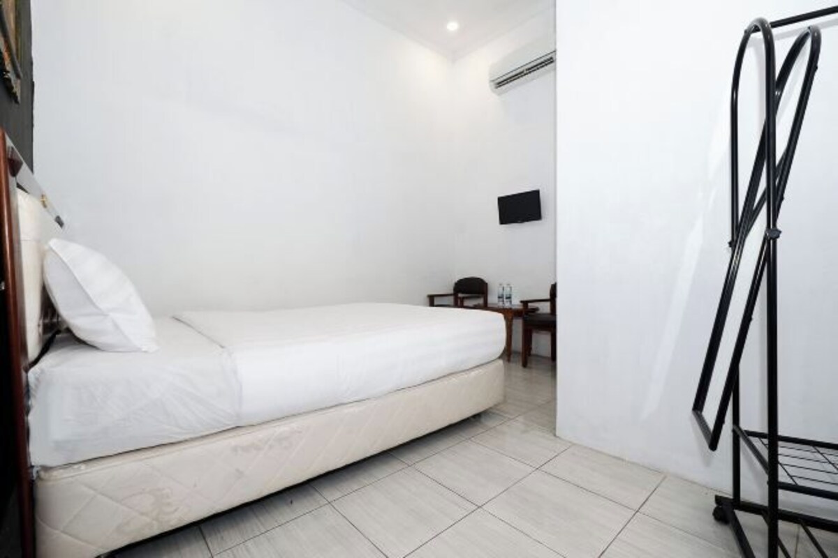 Een enkel bed met een witte matras en kussen, een klein tafeltje met stoel, een aan de muur bevestigde televisie en een metalen rek.