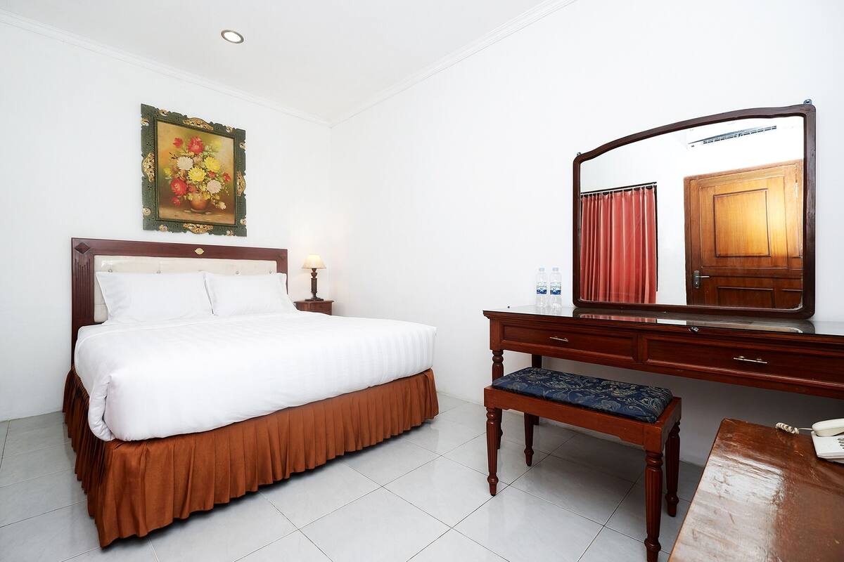 Een hotelkamer met een bed, een houten dressoir, een bankje, een spiegel en een schilderij aan de muur.