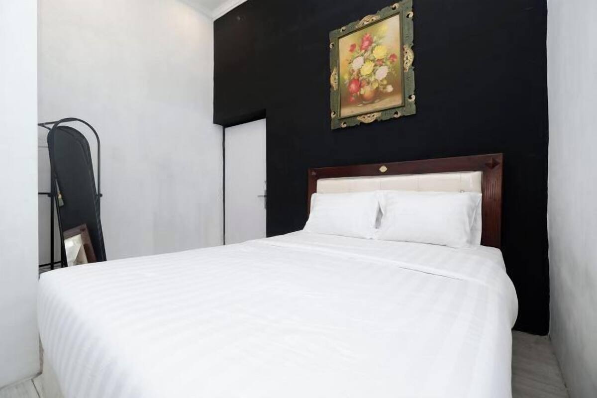 Een net opgemaakt bed met witte lakens, een houten hoofdbord en een lijst met een bloemistisch schilderij erboven.