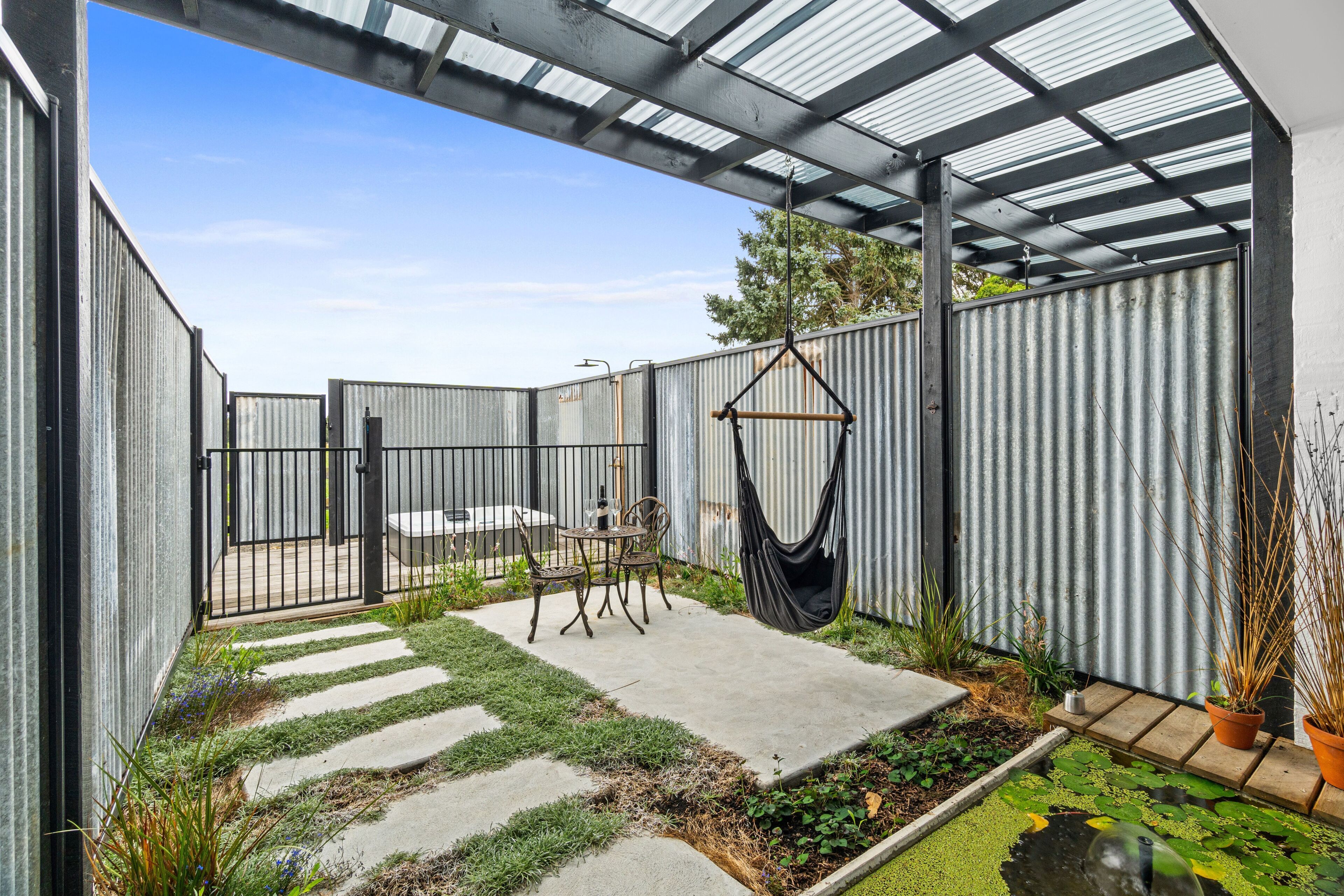 Ein überdachter Patio-Bereich mit einem Betonweg, einem Metallzaun und einem Hängestuhl.