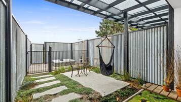 Ein überdachter Patio-Bereich mit einem Betonweg, einem Metallzaun und einem Hängestuhl.