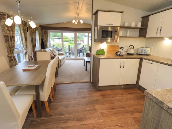 A modern kitchen with a built-in microwave, a toaster, and a kettle on the countertop. There is a dining area with a table and chairs, and a view of a garden through a sliding glass door.