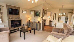A living room with a TV, sofa, coffee table, and a kitchen area in the background. - 41, Grandma's Cottage (Carnforth)