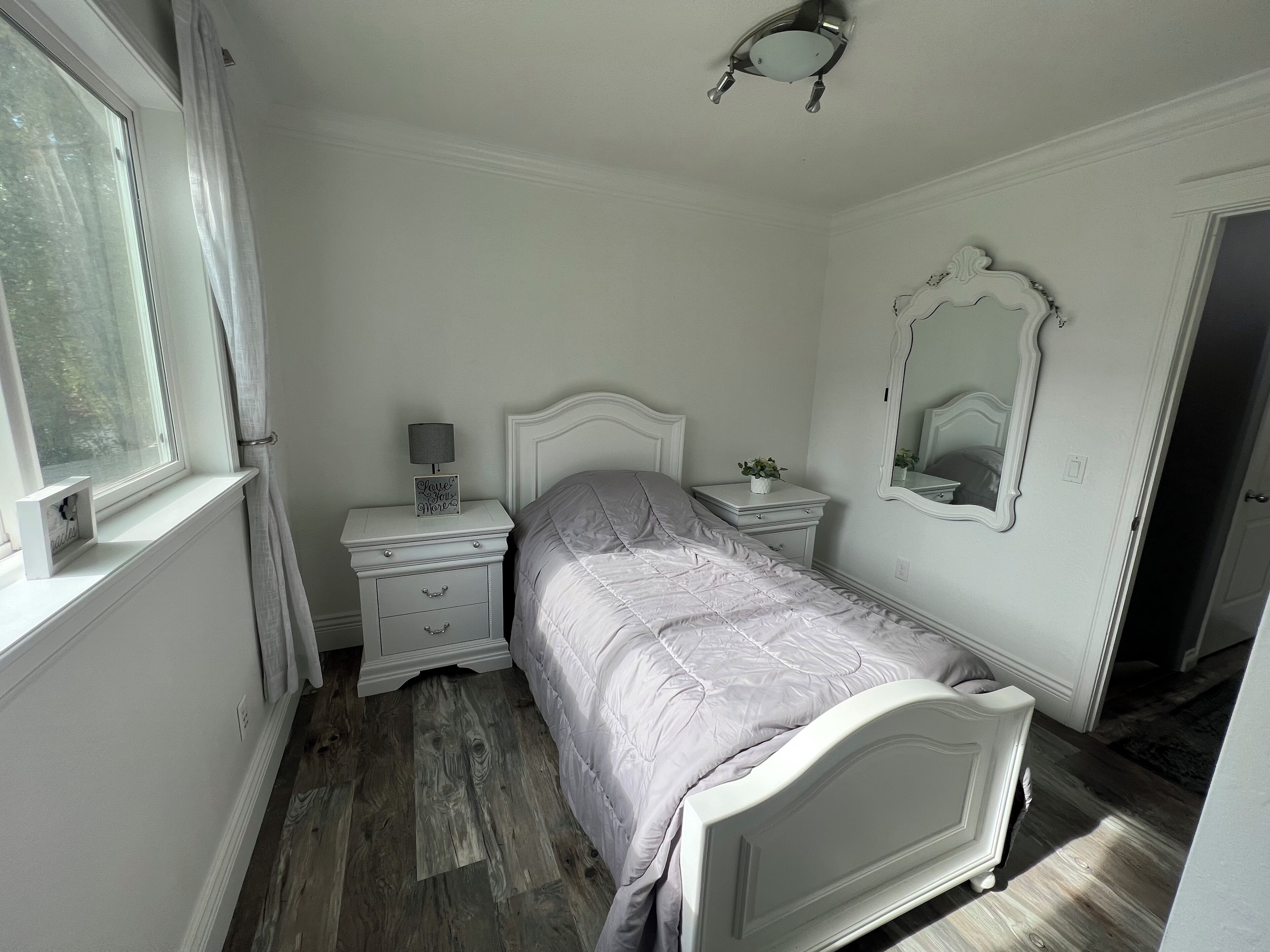 Een slaapkamer met een bed, een nachtkastje met een lamp, een dressoir, een spiegel en een raam met gordijnen.