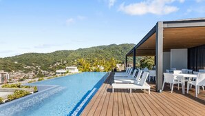 Piscina infinita no terraço com deck de madeira e cadeiras de lounge brancas, com vista para uma paisagem acidentada.