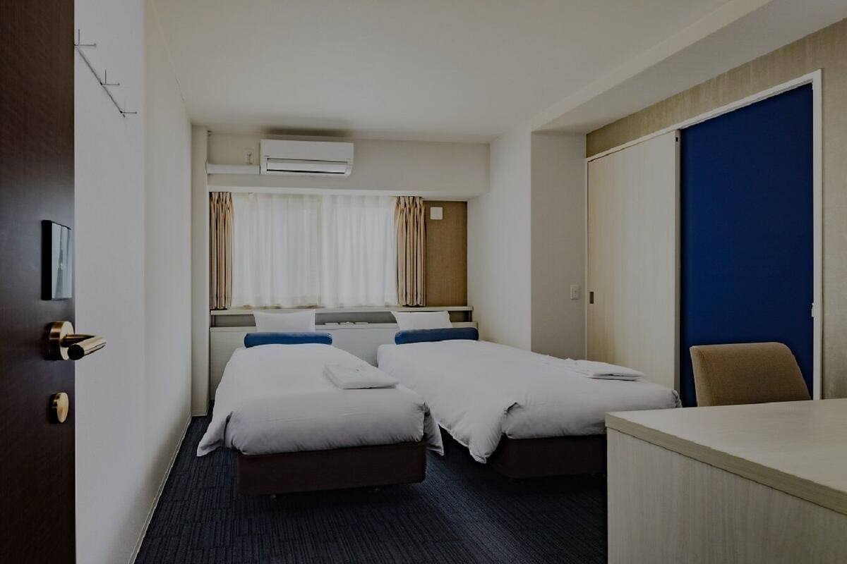 Ein Hotelzimmer mit zwei Betten, einem Schreibtisch, einem Stuhl, einer Klimaanlage und einem mit Vorhängen versehenen Fenster.