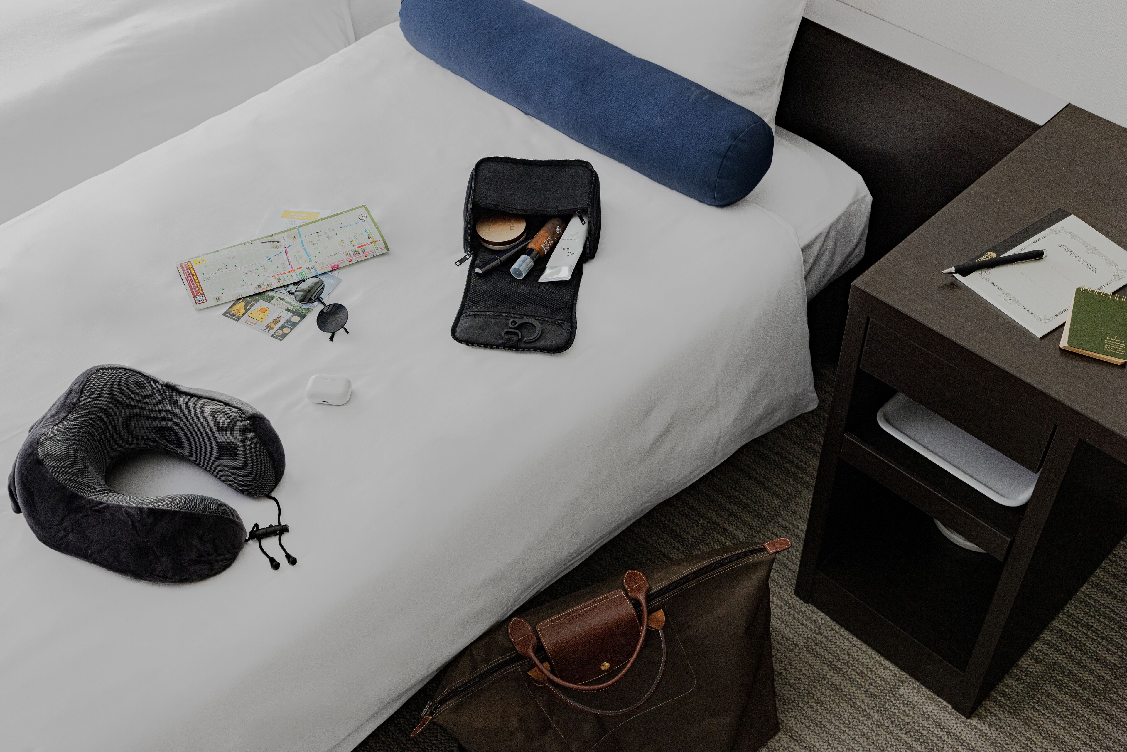 A neatly made bed with a blue pillow, a neck pillow, a travel bag, and a small table with a notebook and pen.