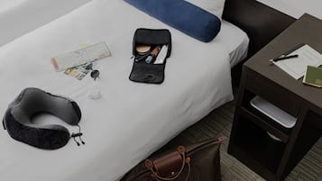 A neatly made bed with a blue pillow, a neck pillow, a travel bag, and a small table with a notebook and pen.