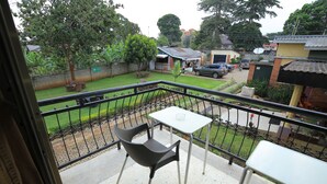 Un balcón con dos sillas y una mesita, con vista a un patio césped que incluye una casa y coches estacionados al fondo.