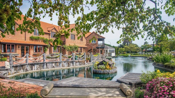 A riverside view with a stone bench, brick buildings, and a small island with plants. - Monaco Resort Apartment with Access to Swimming Pool and Gym Facilities (Nelson)