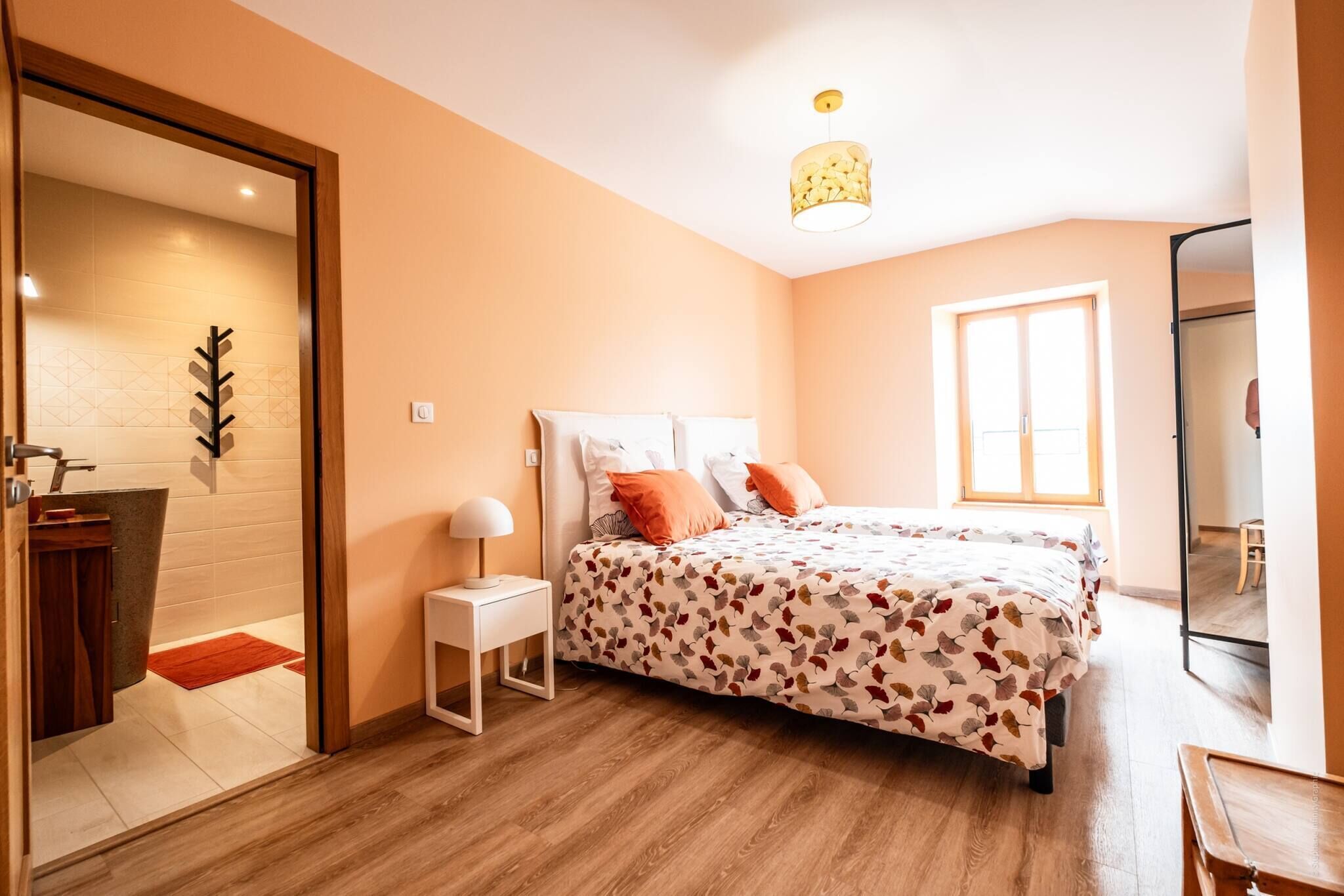 A bedroom with a bed, a nightstand, a lamp, a window, and a door leading to a bathroom.