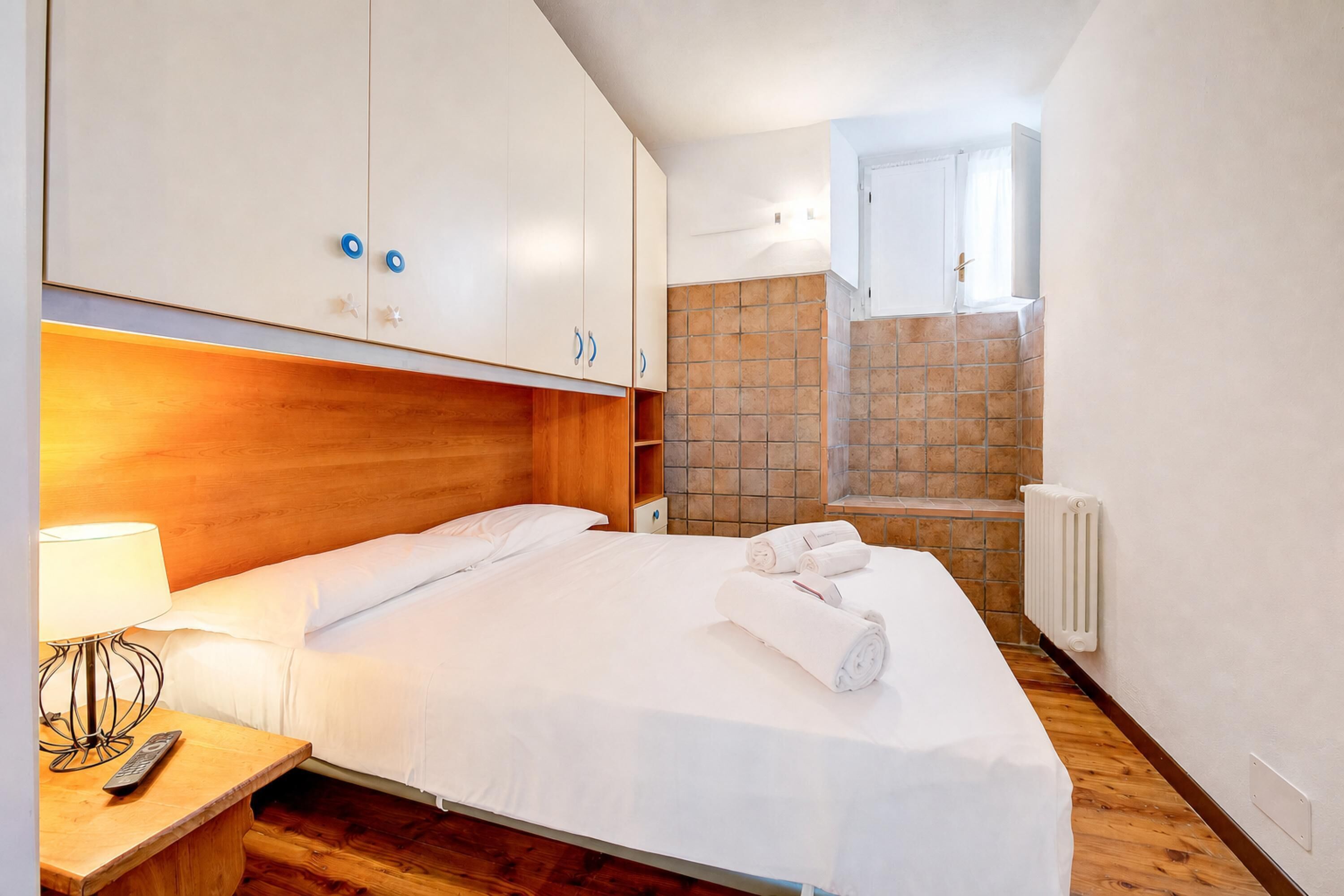 A hotel room with a bed, wooden headboard, white linens, a nightstand with a lamp, a remote control, and a bathroom with a tiled shower area.