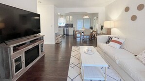 Una sala de estar moderna con televisor de pantalla plana, ventilador de techo, comedor con mesa y sillas, y cocina al fondo.