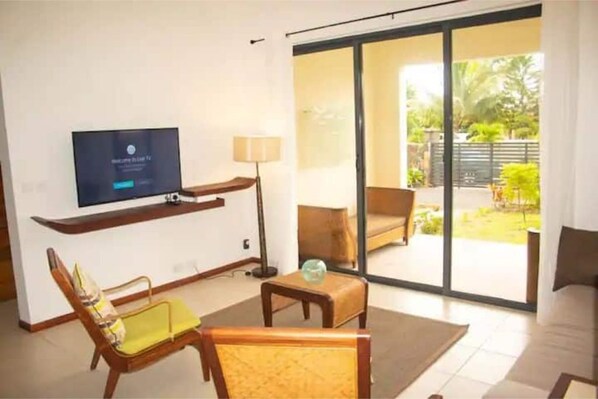 A modern living room with a flat-screen TV, a sofa, a chair, a small table, and a sliding glass door leading to an outdoor area with a view of greenery.