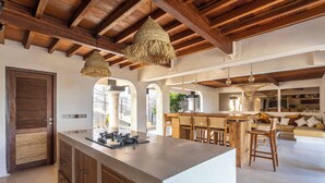 A spacious kitchen with a large island, wooden ceiling beams, and hanging woven light fixtures.