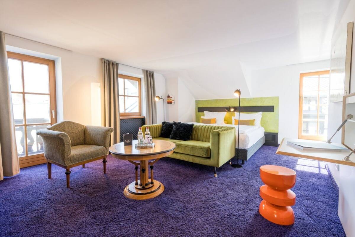 Habitación de hotel con un sofá verde, un sillón de madera, una mesa redonda con bandeja y un taburete naranja brillante.