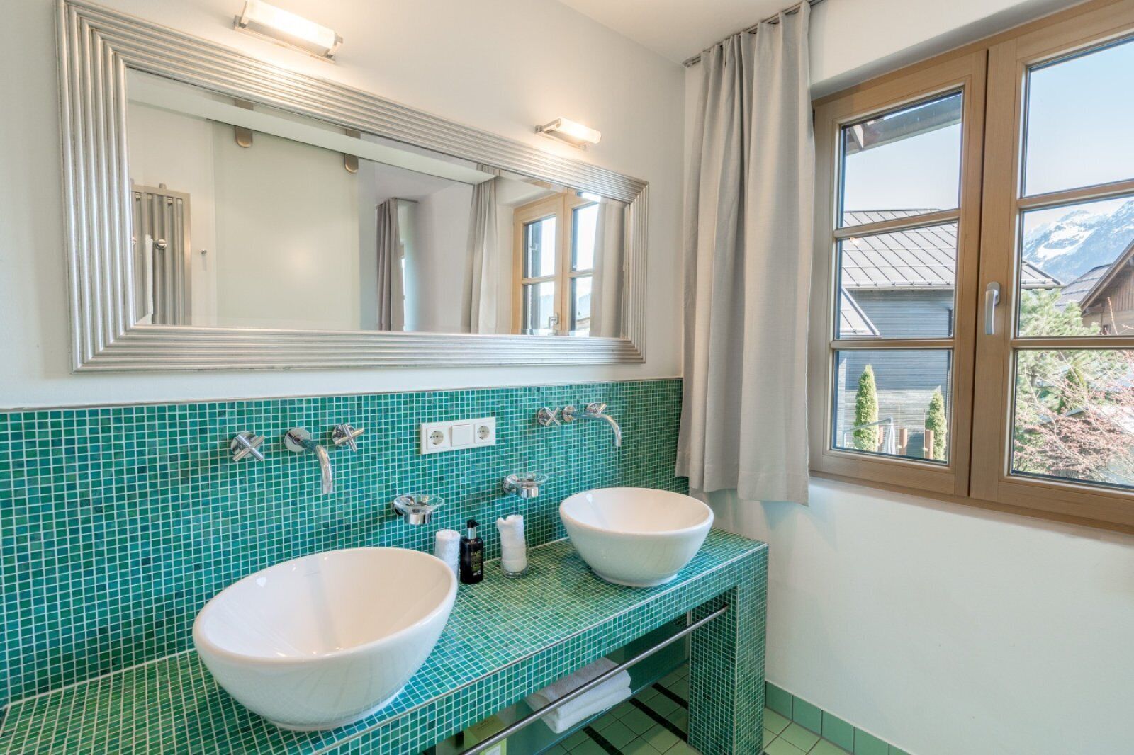 Une salle de bain avec deux lavabos, un grand miroir et une fenêtre donnant sur des immeubles et des arbres.