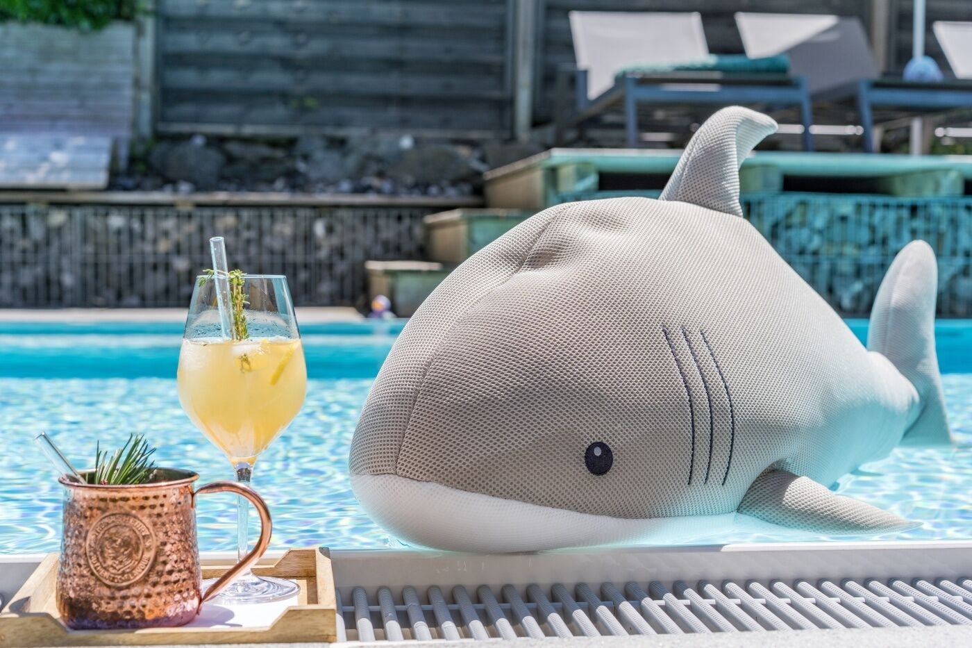 Une scène au bord de la piscine avec un coussin en forme de requin, un verre de limonade et une tasse décorative.